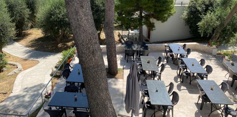 Terrazza con vista mare, tavoli all'aperto tra alberi e fiori.