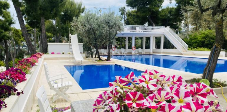 Piscina con scivolo, fiori colorati e sedie bianche, circondata da alberi.